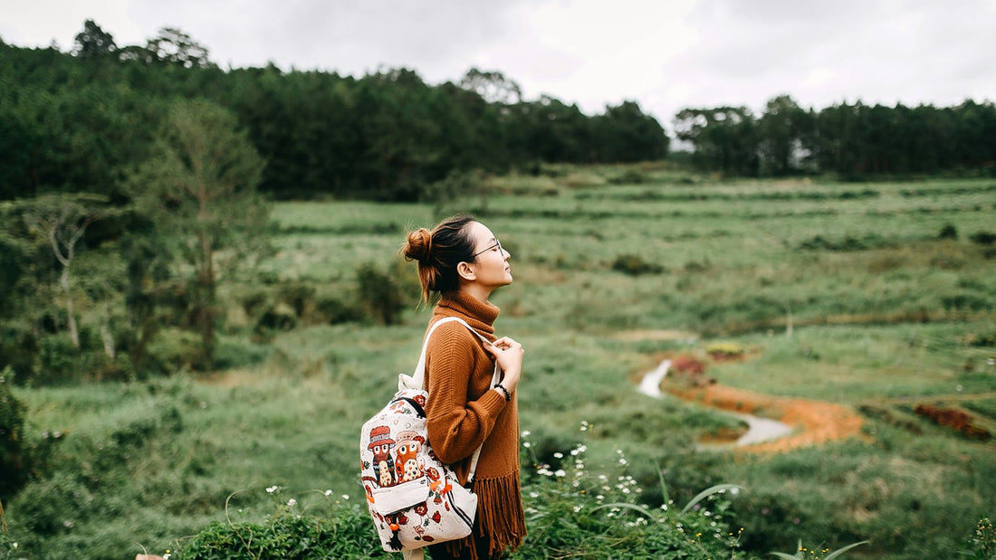 A woman with a backpack pauses on a hillside, looking out over a green valley mid-walk