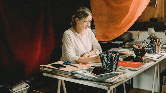 A woman sketches a detailed feather drawing at her desk, fully absorbed in the page