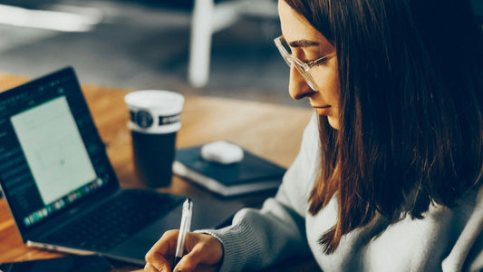 A woman writes in a notebook at a cafe table beside her laptop and a cup of coffee