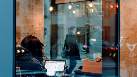 Woman at a café desk glancing between her laptop and phone, attention pulled in two directions, the everyday scatter that makes deep work hard
