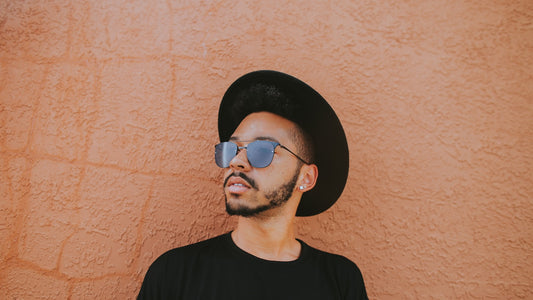 Man in a black hat and sunglasses standing tall against a peach wall, easy self-assurance