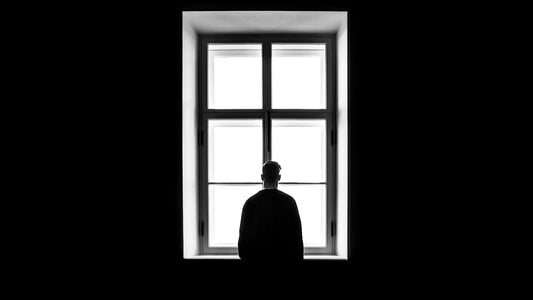 A man stands alone at a window in quiet thought, sitting with the strain of never feeling his achievements are enough