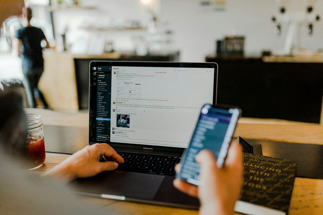 Person split between a laptop and a phone at a café counter, attention divided, the out of sight out of mind pull of ADHD