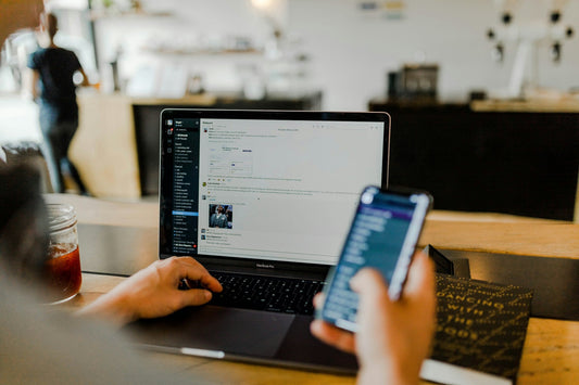 Person split between a laptop and a phone at a café counter, attention divided, the out of sight out of mind pull of ADHD