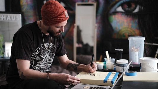 Man in a beanie writing his goals on a pad at a studio desk, colourful mural wall behind him