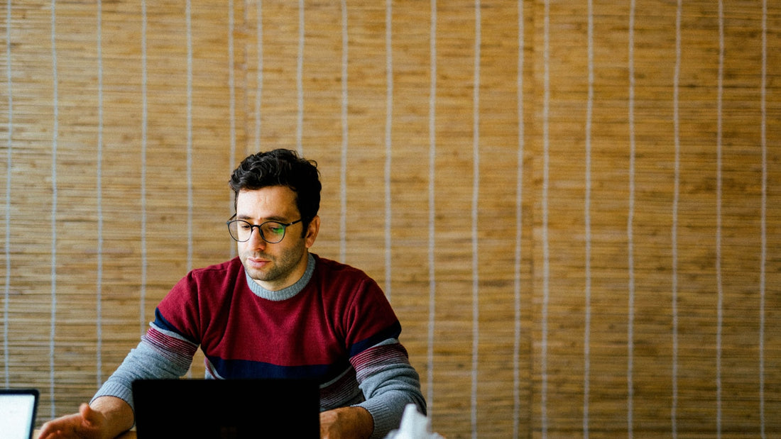 Man with glasses prioritising his day on a laptop at a calm desk, warm bamboo wall behind him