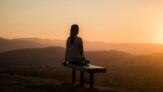 Person sitting alone on a hillside bench at sunset, contemplative, building a positive mindset