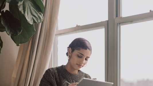 Woman writing in a wellness journal by a window with a laptop nearby, calm and focused