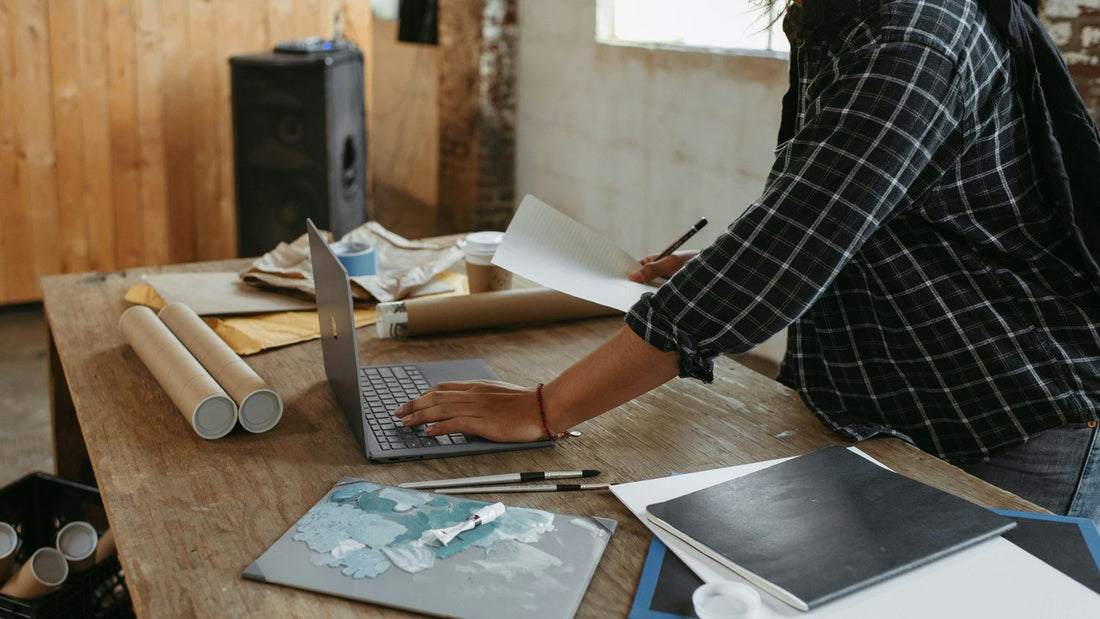Woman in a plaid shirt multitasking between a laptop and scattered papers in a busy studio