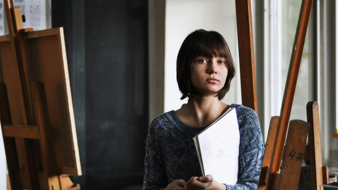 Young woman sitting quietly among art easels holding a sketchbook, looking reflective and self-aware