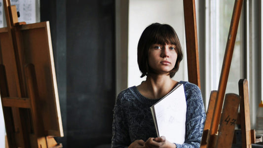 Young woman sitting quietly among art easels holding a sketchbook, looking reflective and self-aware