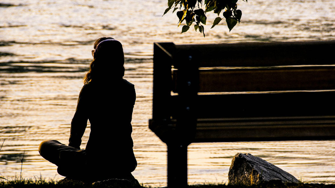 The silhouette of a woman sits cross-legged by the water, meditating quietly at golden hour
