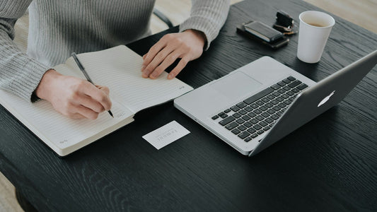 Hands writing plans in a notebook beside a laptop at a minimal desk, elimination planning method